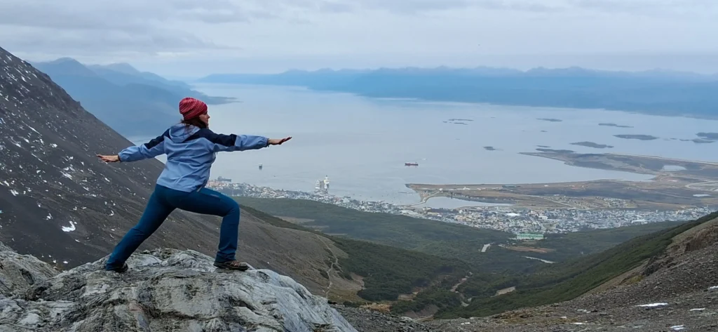 "Mujer realizando la postura del árbol de yoga con botas de trekking en la cima de un cerro al atardecer."